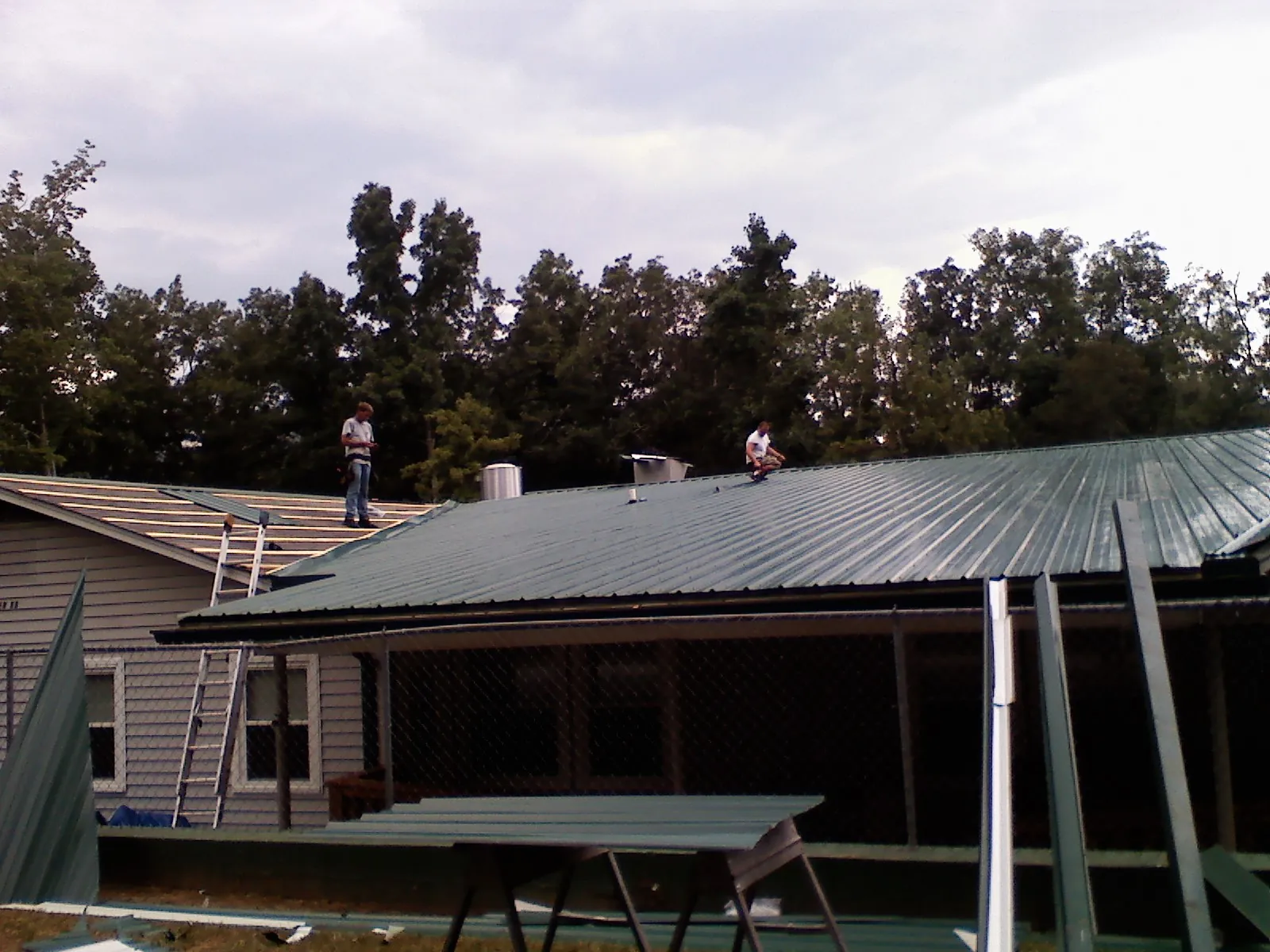 Two roofers installing green metal panels above a covered porch in Forestville